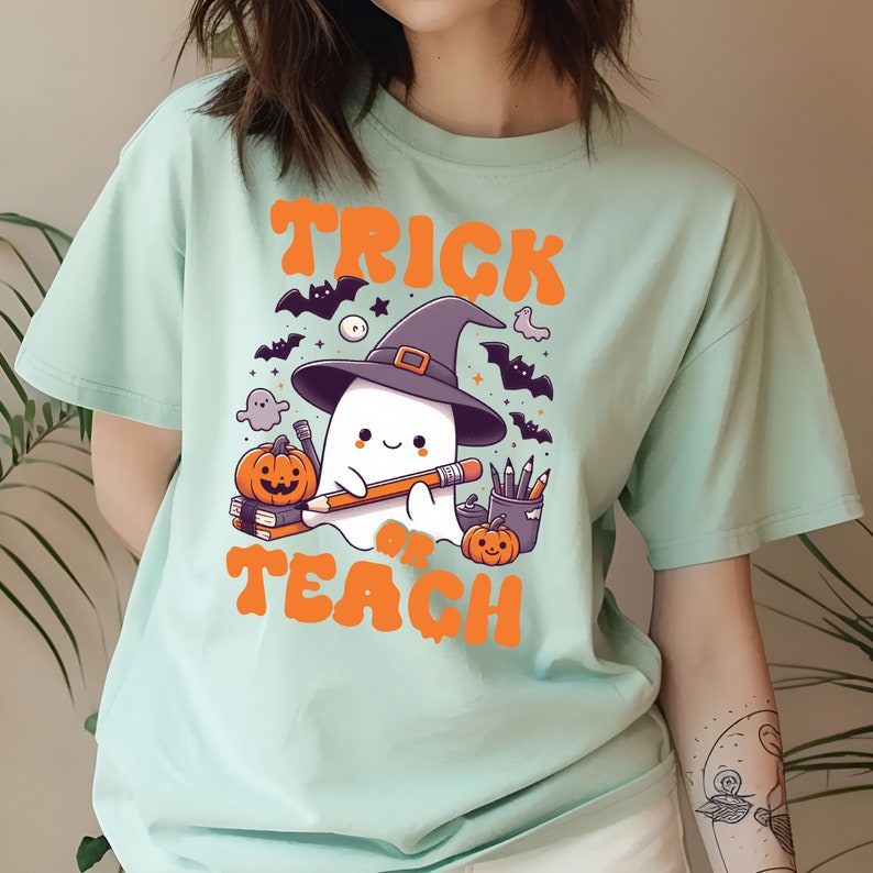 May include: A light blue t-shirt with a cute ghost wearing a witch hat and holding a pencil. The ghost is surrounded by bats, stars, and pumpkins. The text "Trick or Teach" is printed in orange on the shirt.