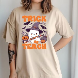 May include: A beige t-shirt with a graphic of a ghost wearing a witch hat and holding a pencil. The text "Trick or Teach" is printed in orange below the ghost.