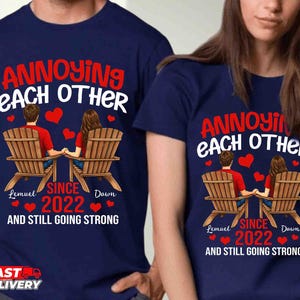 May include: A navy blue t-shirt featuring a graphic of two people sitting in wooden chairs holding hands. The text reads: ANNOYING EACH OTHER SINCE 2022 AND STILL GOING STRONG, with the names Lemuel and Dawn printed below the figures and red hearts.