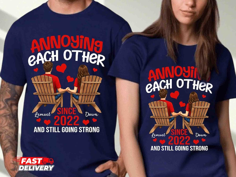 May include: A navy blue t-shirt featuring a graphic of two people sitting in wooden chairs holding hands. The text reads: ANNOYING EACH OTHER SINCE 2022 AND STILL GOING STRONG, with the names Lemuel and Dawn printed below the figures and red hearts.