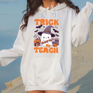 May include: White hoodie with a graphic of a ghost wearing a witch hat and holding a pencil. The text "Trick or Teach" is printed below the ghost.