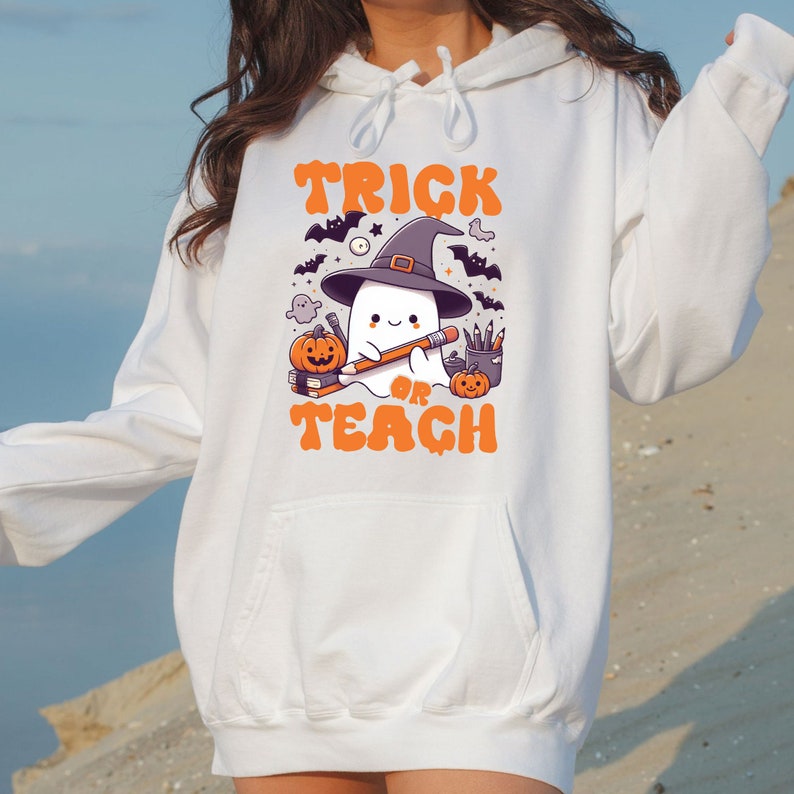 May include: White hoodie with a graphic of a ghost wearing a witch hat and holding a pencil. The text "Trick or Teach" is printed below the ghost.