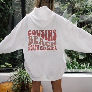 May include: White hooded sweatshirt with the text "COUSINS BEACH NORTH CAROLINA" printed on the back in pink and red retro lettering.