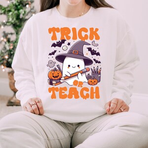 May include: White sweatshirt with a cute ghost wearing a witch hat and holding a pencil. The text "Trick or Teach" is printed in orange on the front of the sweatshirt.