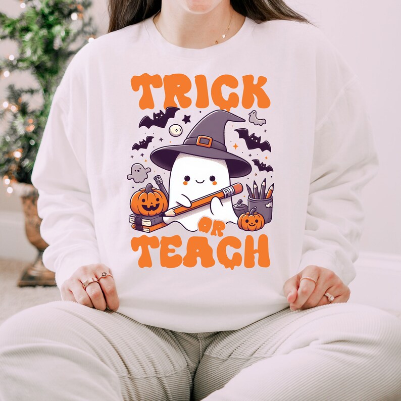May include: White sweatshirt with a cute ghost wearing a witch hat and holding a pencil. The text "Trick or Teach" is printed in orange on the front of the sweatshirt.