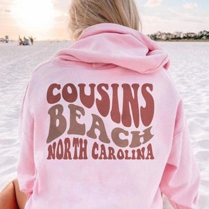 May include: Pink hooded sweatshirt with the text "COUSINS BEACH NORTH CAROLINA" in a retro font.