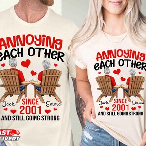 May include: Two cream-colored t-shirts featuring a graphic of two people sitting in wooden chairs holding hands. The text reads: ANNOYING EACH OTHER SINCE 2001 AND STILL GOING STRONG, with the names Jack and Emma printed below the chairs.