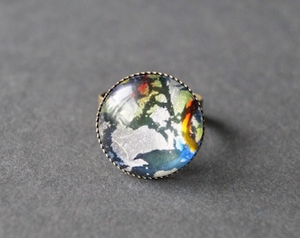 Pode incluir: Um anel com um design abstrato redondo, prateado, azul, verde e amarelo, engastado em uma moldura de metal dourado.