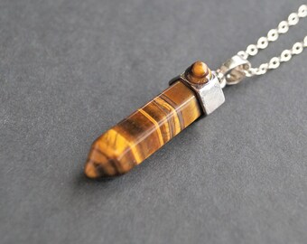 May include: A pendant necklace featuring a polished tiger's eye gemstone. The pendant is a brown and gold hexagonal shape, suspended from a delicate silver chain. The gemstone has natural stripes and is set in a silver metal cap.