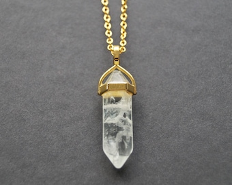 Puede incluir: Un collar de cadena dorada con un colgante de cristal de cuarzo transparente. El cristal tiene forma de punta y está engastado en un bisel dorado.