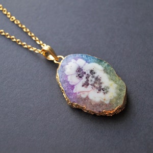 Peut inclure: Un collier en cha&icirc;ne dor&eacute;e avec un pendentif en forme de tranche d'agate arc-en-ciel avec un motif de fleur blanche.