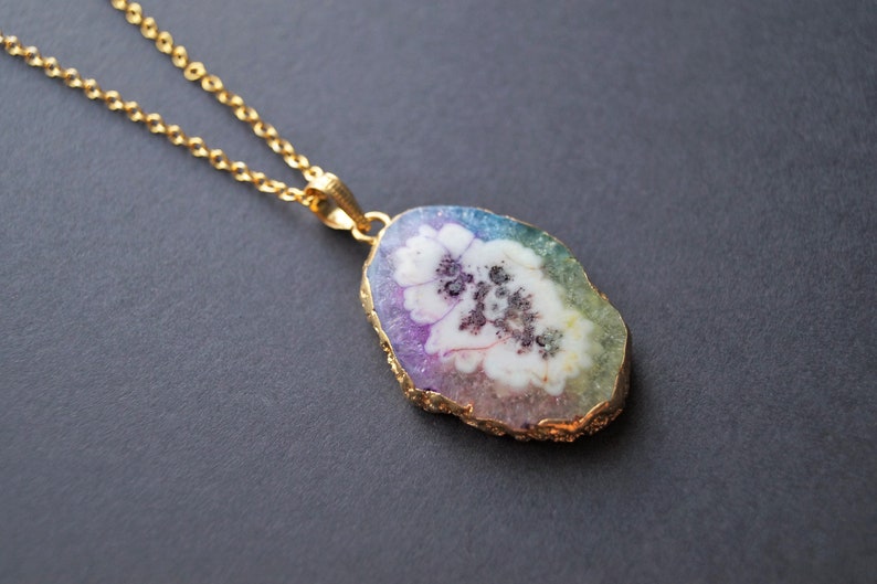 Peut inclure: Un collier en cha&icirc;ne dor&eacute;e avec un pendentif en forme de tranche d'agate arc-en-ciel avec un motif de fleur blanche.