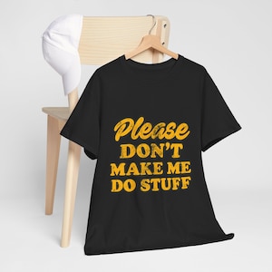 May include: A black short-sleeved t-shirt featuring the yellow text "Please DON'T MAKE ME DO STUFF" in a distressed font style, displayed alongside a plain white baseball cap hanging on a light-colored wooden chair.