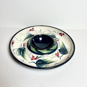 Puede incluir: Una fuente redonda de cerámica blanquecina con un cuenco central para salsas. La fuente está decorada con un borde azul, hojas verdes, flores rojas y enredaderas azules. El cuenco para salsas es azul oscuro con rayas verdes.