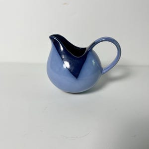 May include: A ceramic pitcher with a rounded body and a curved handle. The pitcher is primarily light blue with a dark blue triangular accent on the front. The interior is dark blue. This decorative pitcher is suitable for serving beverages.