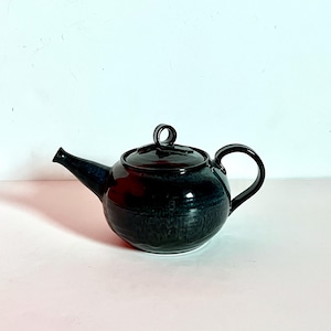 Pode incluir: Uma chaleira de cerâmica azul escura com um corpo arredondado, uma alça curva e um bico. A tampa tem uma alça de laço. A chaleira tem um acabamento brilhante e assenta sobre uma superfície branca. A chaleira foi projetada para preparar chá.