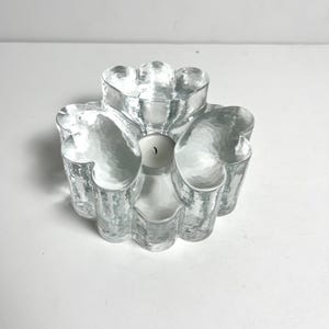 Puede incluir: Un portavelas de vidrio transparente con una superficie texturizada. El portavelas tiene un diseño en forma de flor con seis secciones curvas. Una vela blanca se coloca en el centro del soporte. El portavelas está sobre una superficie blanca.