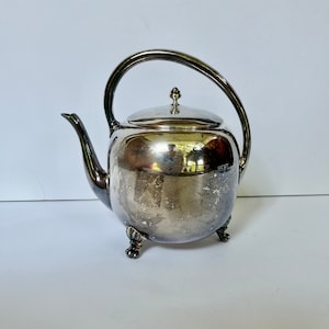 Może przedstawiać: Srebrny czajnik z zaokrąglonym korpusem i trzema nóżkami. Czajnik ma błyszczące wykończenie i lekko matowe wykończenie.