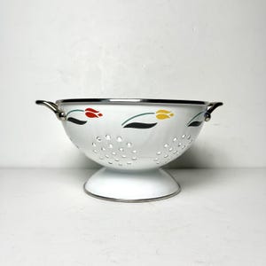 May include: A white enameled metal colander featuring a pattern of stylized red, yellow, and blue tulips with green leaves. The bowl has a pedestal base, two side handles, and a silver-toned rim, with circular drainage holes throughout the body.