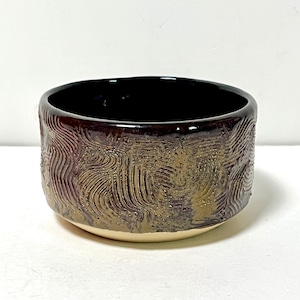 May include: A round ceramic bowl featuring a glossy black interior and a textured exterior with a swirling, raised pattern in metallic bronze and dark purple tones. The base of the bowl is a smooth, unglazed cream color, contrasting with the dark finish.