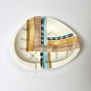 Peut inclure: Cendrier de style moderne du milieu du siècle, de couleur crème, avec une forme asymétrique unique. Le cendrier présente un motif géométrique dans les tons de marron, or, sarcelle et blanc. Le motif comprend des éléments en forme d'éclat d'étoile et linéaires.