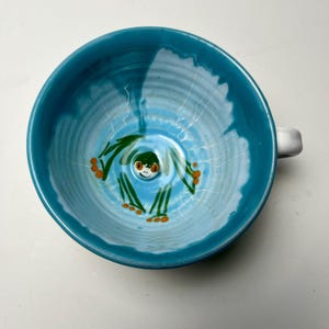 Könnte beinhalten: Eine tealblaue Keramiktasse mit weißem Überzug und einem handgemalten grünen Froschdesign. Der Frosch hat orangefarbene Füße und ein weißes Gesicht mit schwarzen Augen.