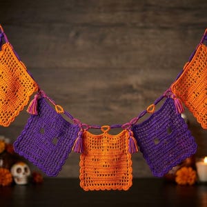 May include: A crochet bunting banner featuring alternating orange and purple square panels with intricate openwork patterns. The banner is accented with small tassels and two white skull beads at the ends, suspended by a matching cord.
