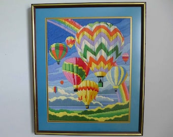 May include: Framed cross-stitch artwork depicting a colorful scene of hot air balloons floating in the sky with a rainbow in the background.