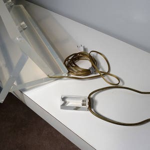 May include: A clear acrylic lamp base with a gold metal harp and cord. The lamp base is folded in half and the cord is coiled.