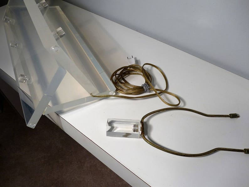 May include: A clear acrylic lamp base with a gold metal harp and cord. The lamp base is folded in half and the cord is coiled.