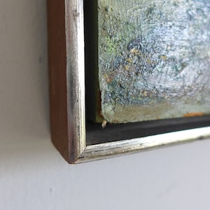 Puede incluir: Primer plano de la esquina de una pintura enmarcada. El marco es de madera con un borde plateado. La pintura es abstracta y presenta tonos de verde, azul y blanco.