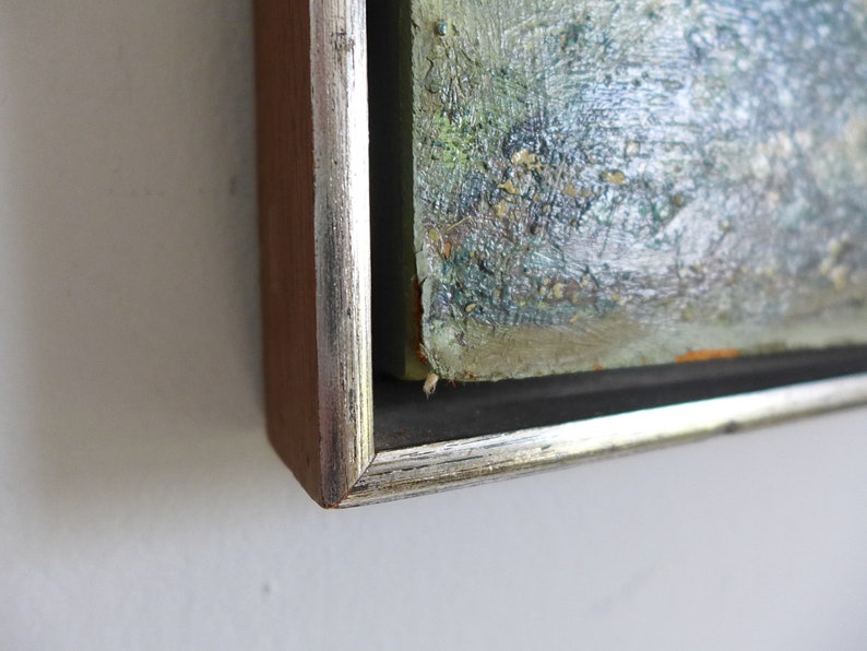 Puede incluir: Primer plano de la esquina de una pintura enmarcada. El marco es de madera con un borde plateado. La pintura es abstracta y presenta tonos de verde, azul y blanco.