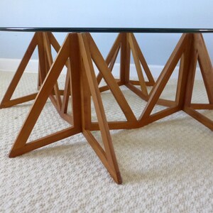 May include: A round glass top coffee table with a unique, geometric wooden base made of triangular shapes. The base is made of light brown wood and the table is sitting on a white rug.