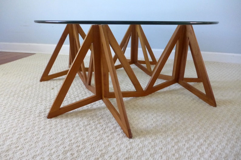 May include: A round glass top coffee table with a unique, geometric wooden base made of triangular shapes. The base is made of light brown wood and the table is sitting on a white rug.