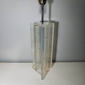 May include: A clear acrylic lamp with a geometric design. The lamp has a metal rod that extends from the base to the top, where a light bulb is attached.