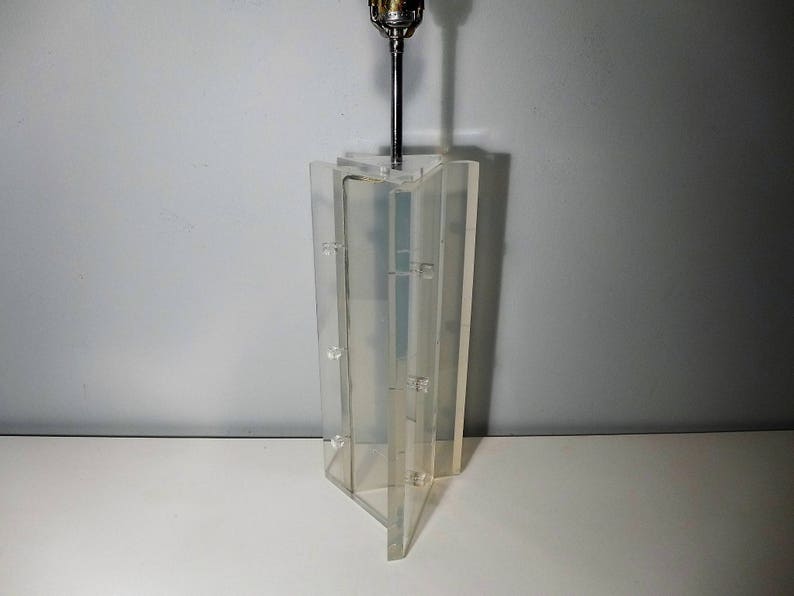 May include: A clear acrylic lamp with a geometric design. The lamp has a metal rod that extends from the base to the top, where a light bulb is attached.