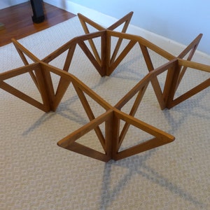 May include: A wooden coffee table base with a geometric design. The base is made of multiple triangular pieces of wood that are joined together to form a square shape. The table base is sitting on a white carpet.