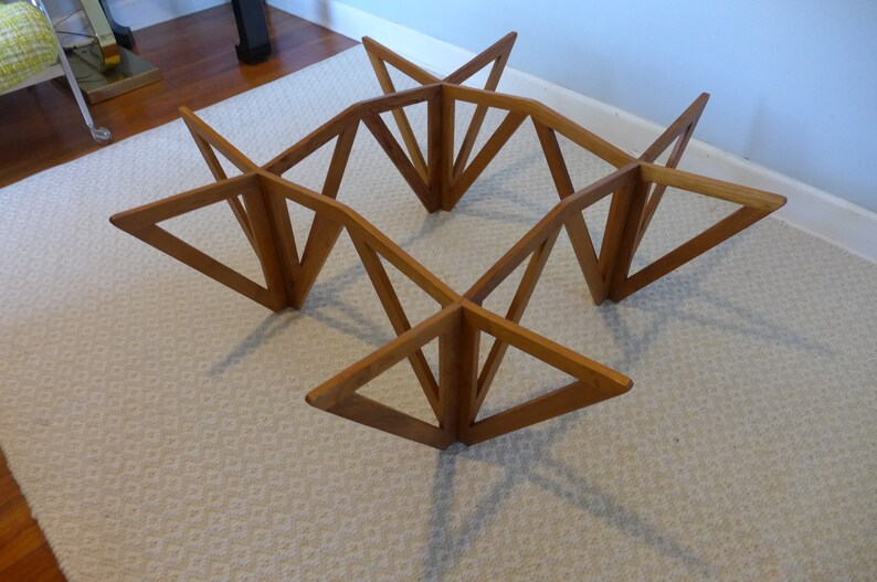 May include: A wooden coffee table base with a geometric design. The base is made of multiple triangular pieces of wood that are joined together to form a square shape. The table base is sitting on a white carpet.