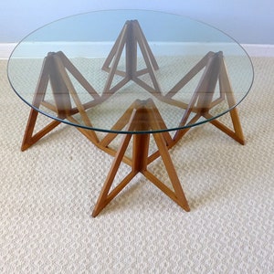 May include: A round glass top coffee table with a unique wooden base made of triangular shapes. The base is made of light brown wood and the table is sitting on a white rug.