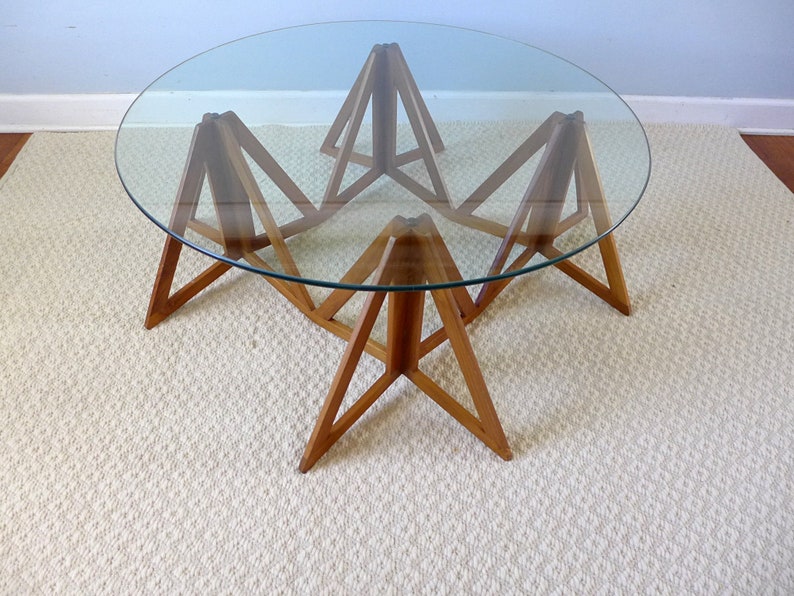 May include: A round glass top coffee table with a unique wooden base made of triangular shapes. The base is made of light brown wood and the table is sitting on a white rug.