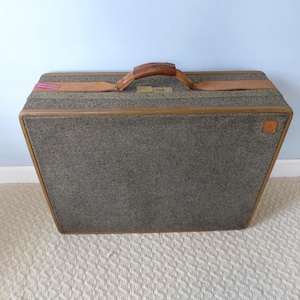 May include: A vintage suitcase with a gray tweed exterior and brown leather handle and trim. The suitcase has a rectangular shape with rounded corners and a small tag on the side.