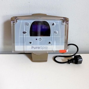 May include: A rectangular PureSpa control panel with a clear protective cover. The panel has a digital display and several control buttons. A black power cord with a plug extends from the bottom right. The panel is mounted on a beige bracket.