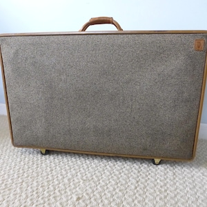 May include: Rectangular vintage suitcase with a tweed fabric front and a brown leather handle. The suitcase has a wooden frame with gold-colored feet and a small logo on the front.