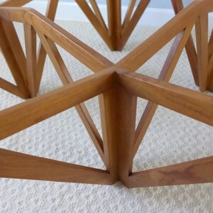 May include: A wooden table base with a geometric design. The base is made of multiple pieces of wood that are joined together to form a star shape. The table base is a light brown color.