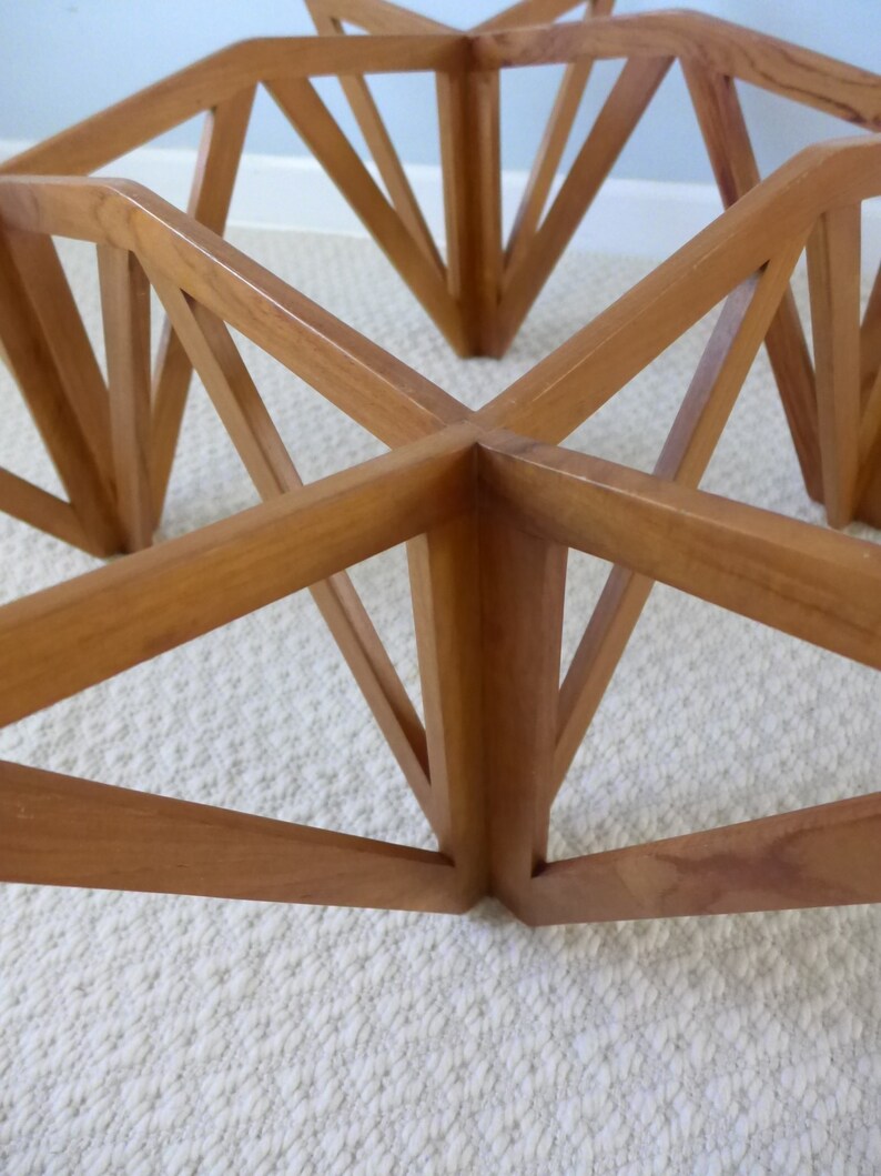May include: A wooden table base with a geometric design. The base is made of multiple pieces of wood that are joined together to form a star shape. The table base is a light brown color.