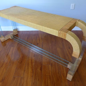 May include: A long, rectangular console table with a woven top and clear glass legs. The table has a curved design and is made of natural materials.