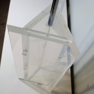 May include: A clear acrylic prism with a metal rod inserted through the top. The prism is made of multiple pieces of acrylic that are joined together. The prism is sitting on a white surface.