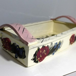 May include: Cream-colored rectangular wooden box with a pink handle and painted floral designs in red, blue, and black. The handle has a crosshatch pattern. The box is a decorative storage container.