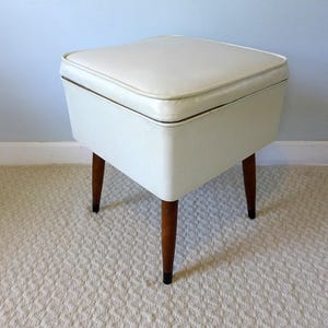 May include: A vintage white vinyl ottoman with a square top and four tapered wooden legs. The ottoman has a cream-colored cushion and a contrasting dark trim. The legs are a dark brown color.
