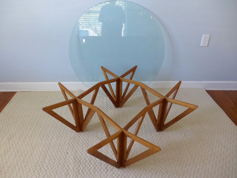 May include: A mid-century modern coffee table with a round glass top and a geometric wooden base. The base is made of four sets of triangular pieces that intersect to form a star shape.